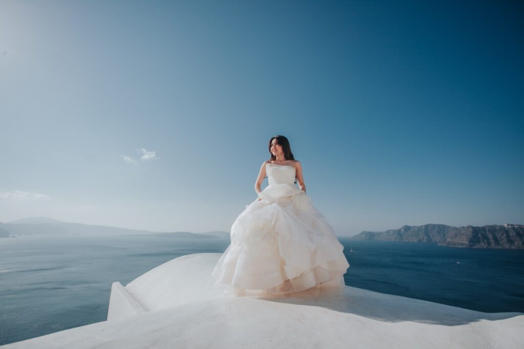 Irene stands in a full white ball gown on a Santorini rooftop over the sea