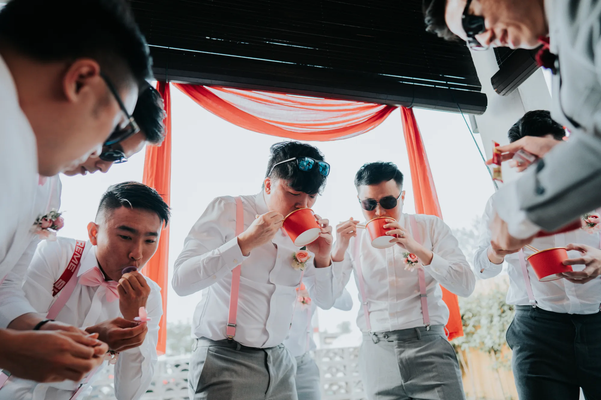 A group of men in formal attire partakes in a ceremonial drink at an outdoor event under bright daylight.