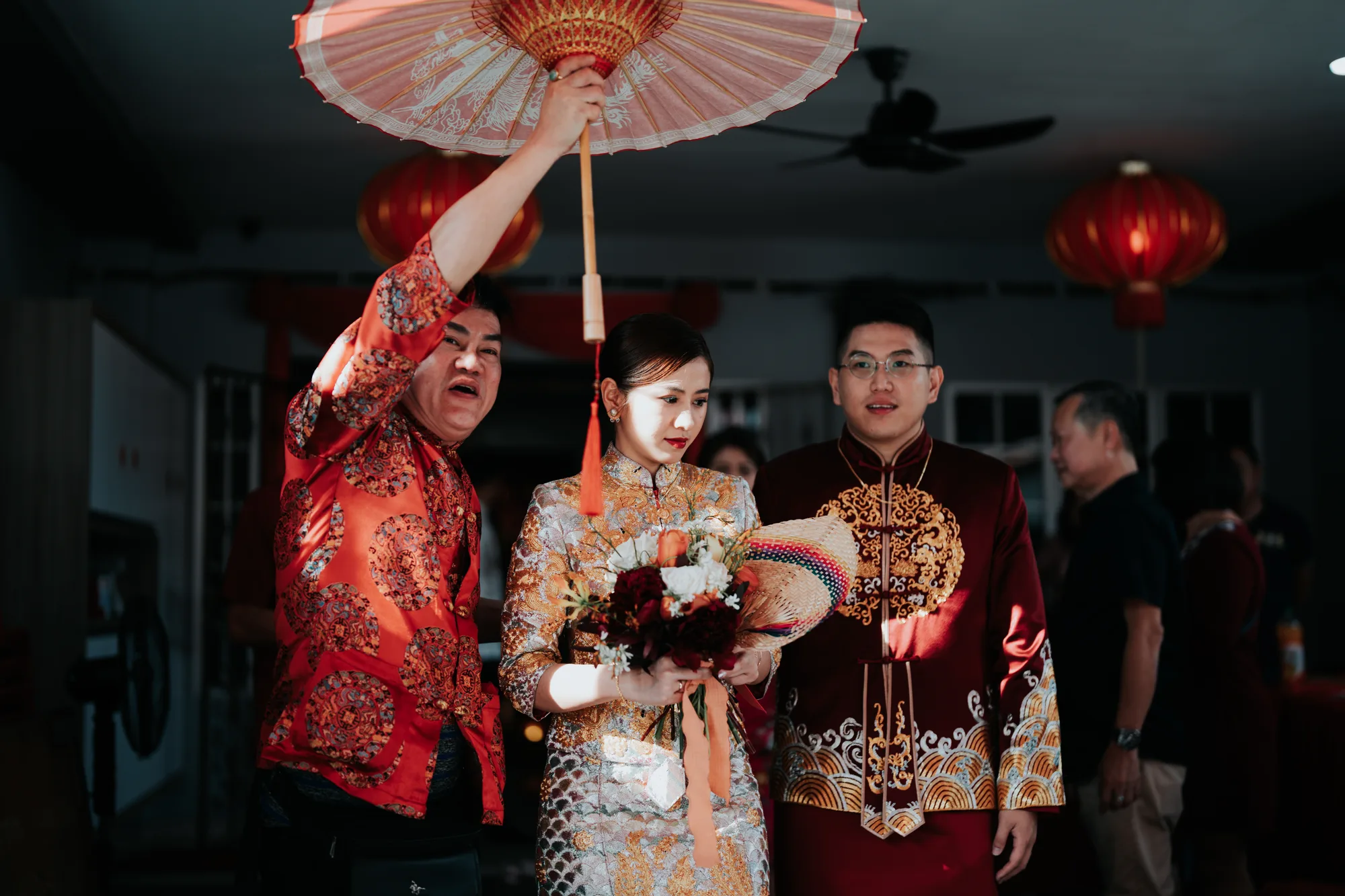 A bride and groom in traditional attire participate in a wedding ceremony under an ornate parasol held by another individual