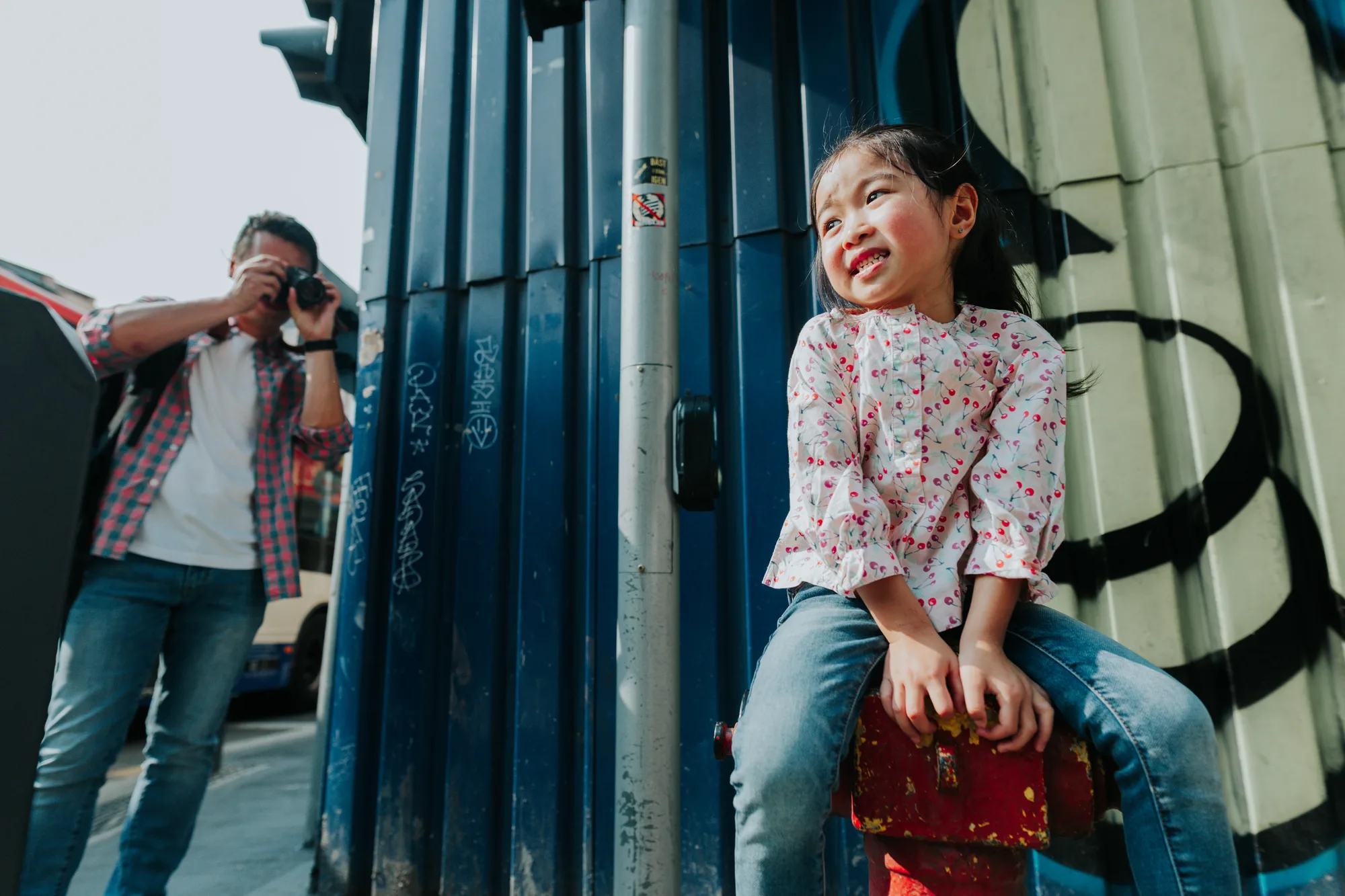 A young girl sits on a red suitcase against a graffiti wall while her father photographs her