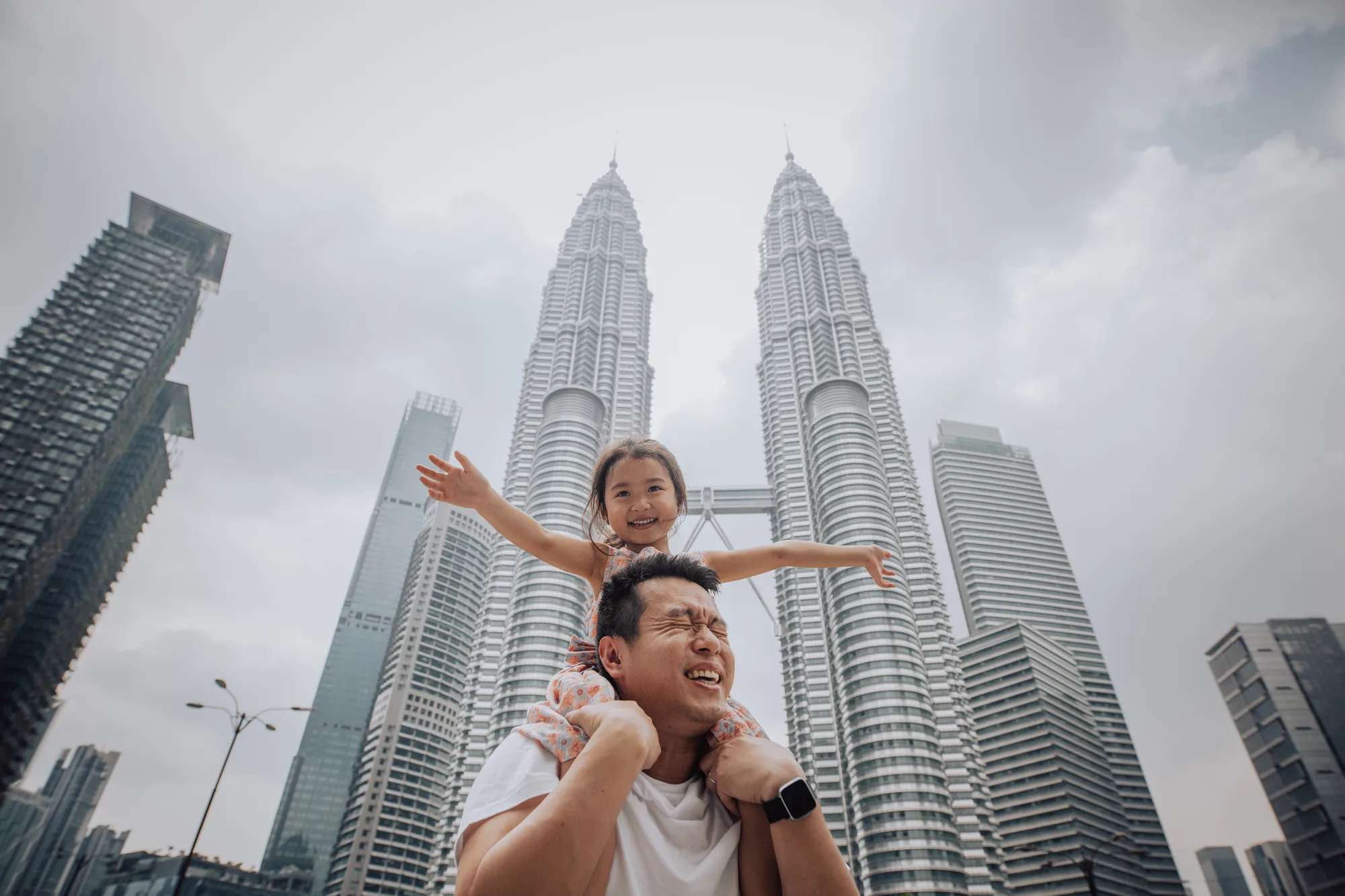 A father carries his daughter on his shoulders in front of the Petronas Twin Towers