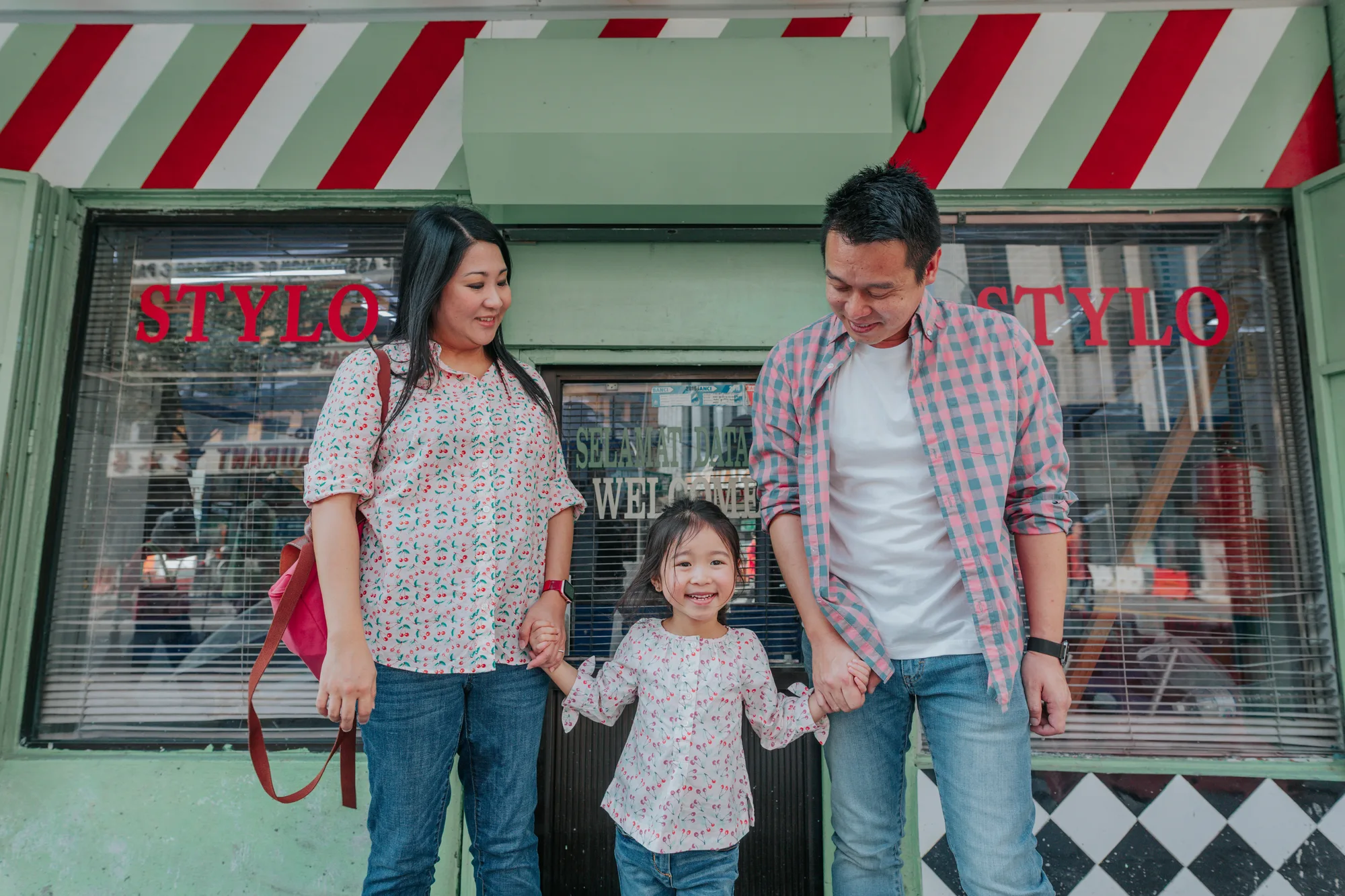 A family of three in matching red and white stands in front of a shop with a striped awning