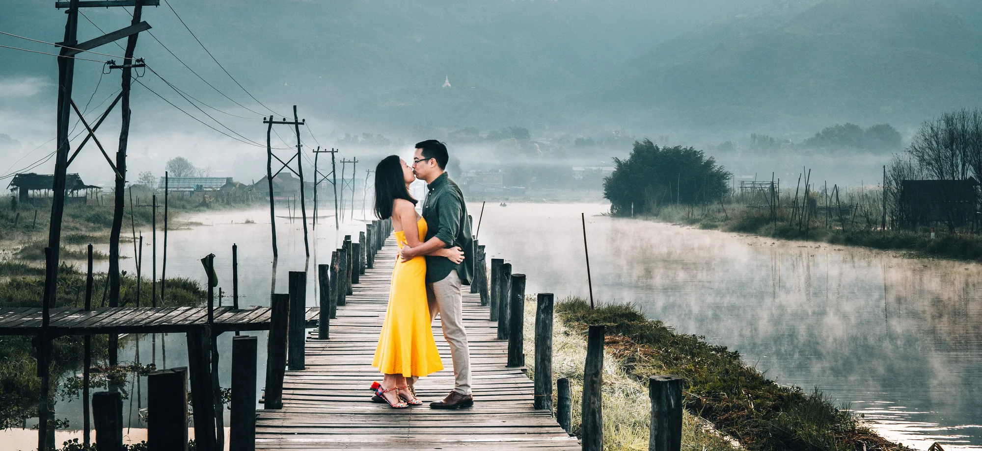A couple shares a tender moment on a wooden bridge over a misty river, bathed in soft morning light.