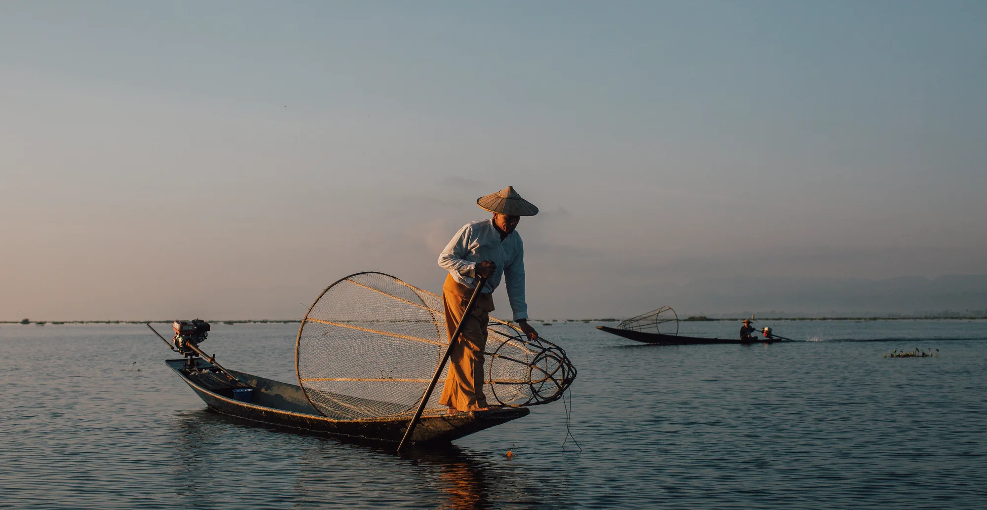 A fisherman in traditional attire stands on a boat holding a circular fishing net at sunset