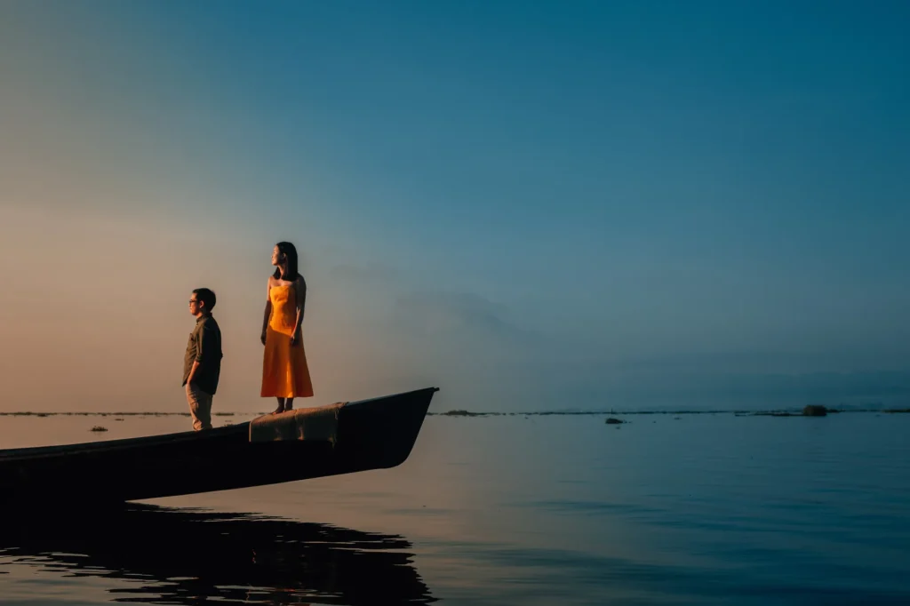 Two people stand on a boat at sunset, silhouetted against the sky and water