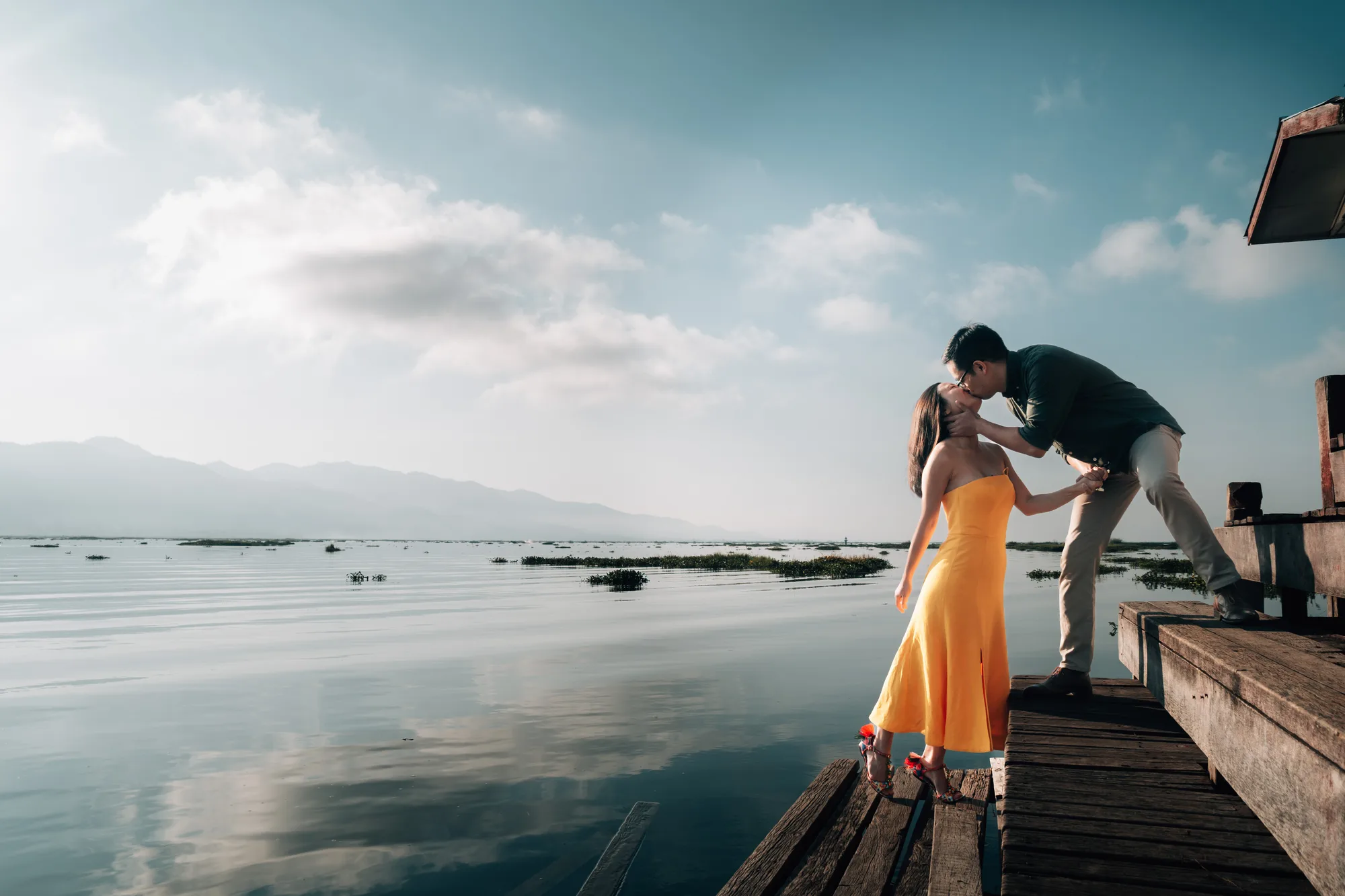 A couple shares a tender moment on a wooden dock by the serene lake under a soft blue sky with scattered clouds.