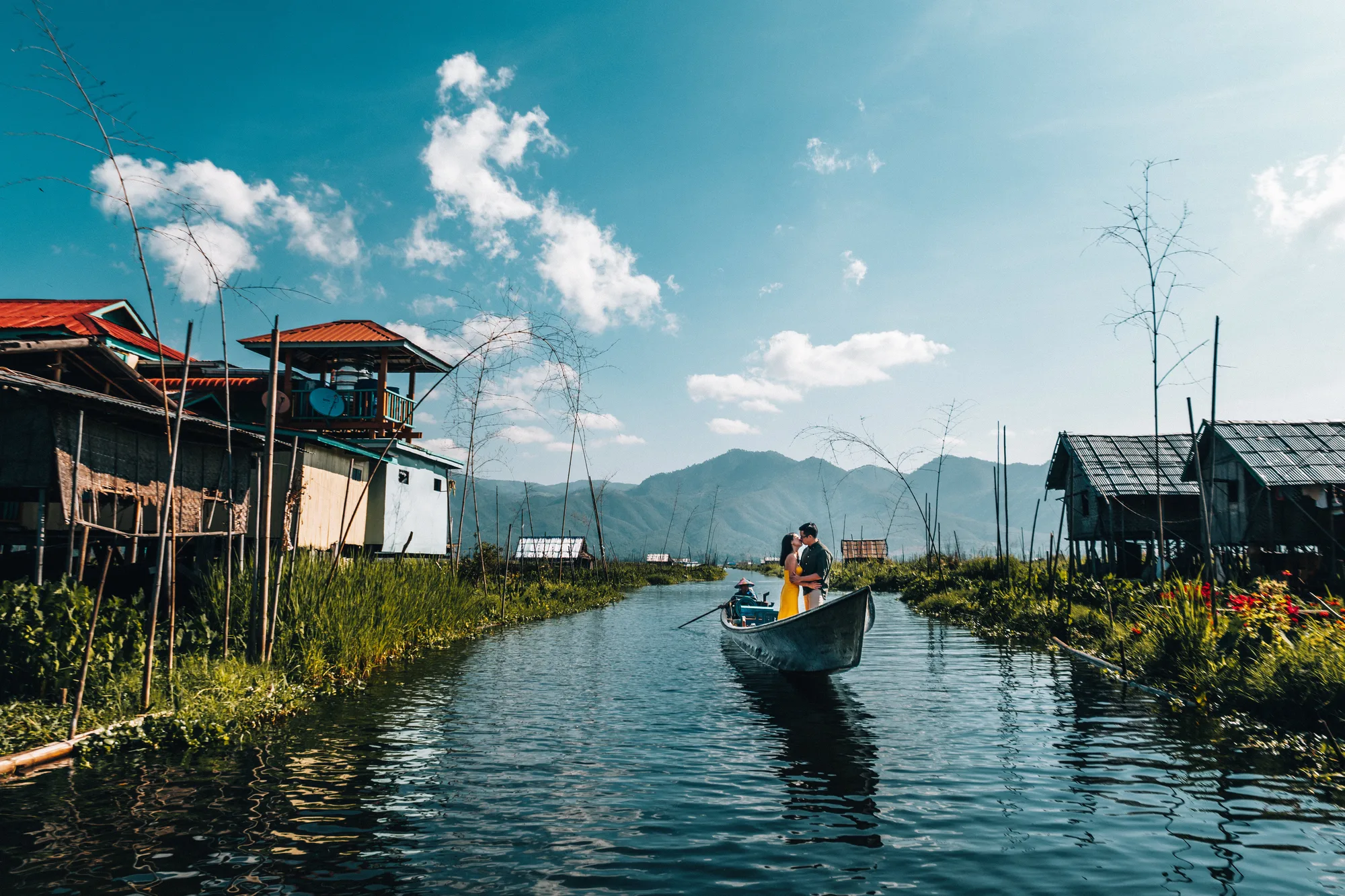 A couple shares a tender moment on a boat amidst serene waters and traditional stilt houses under a bright blue sky with scat