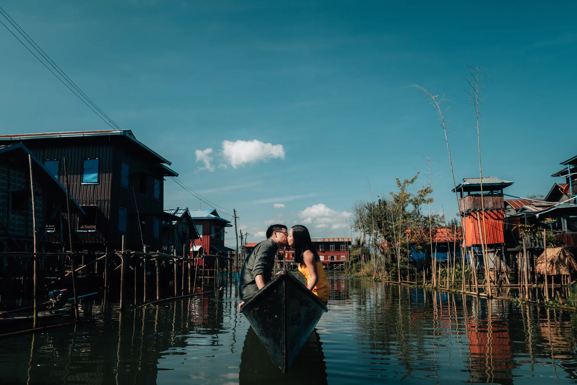 A couple shares a tender moment in a small boat amidst a serene village setting under clear blue skies.
