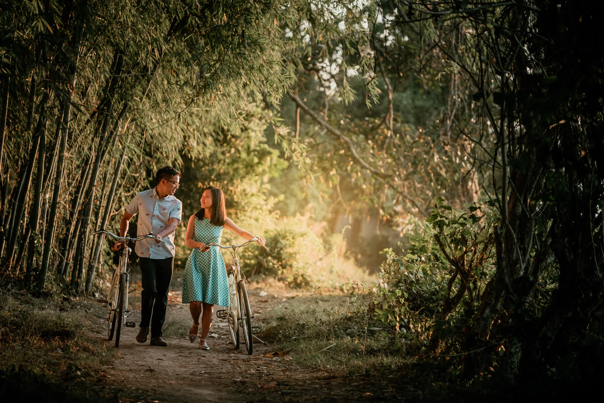 A couple walks with bicycles through a sunlit forest path, creating a serene and intimate moment.
