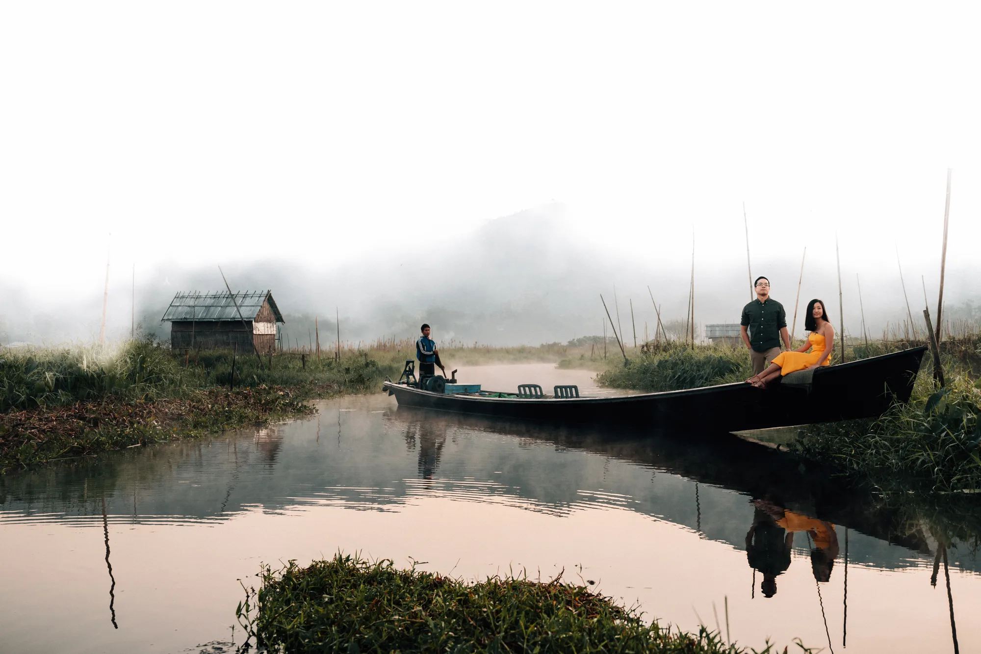 A man and a woman sit in a boat on a misty riverbank with a small hut in the background