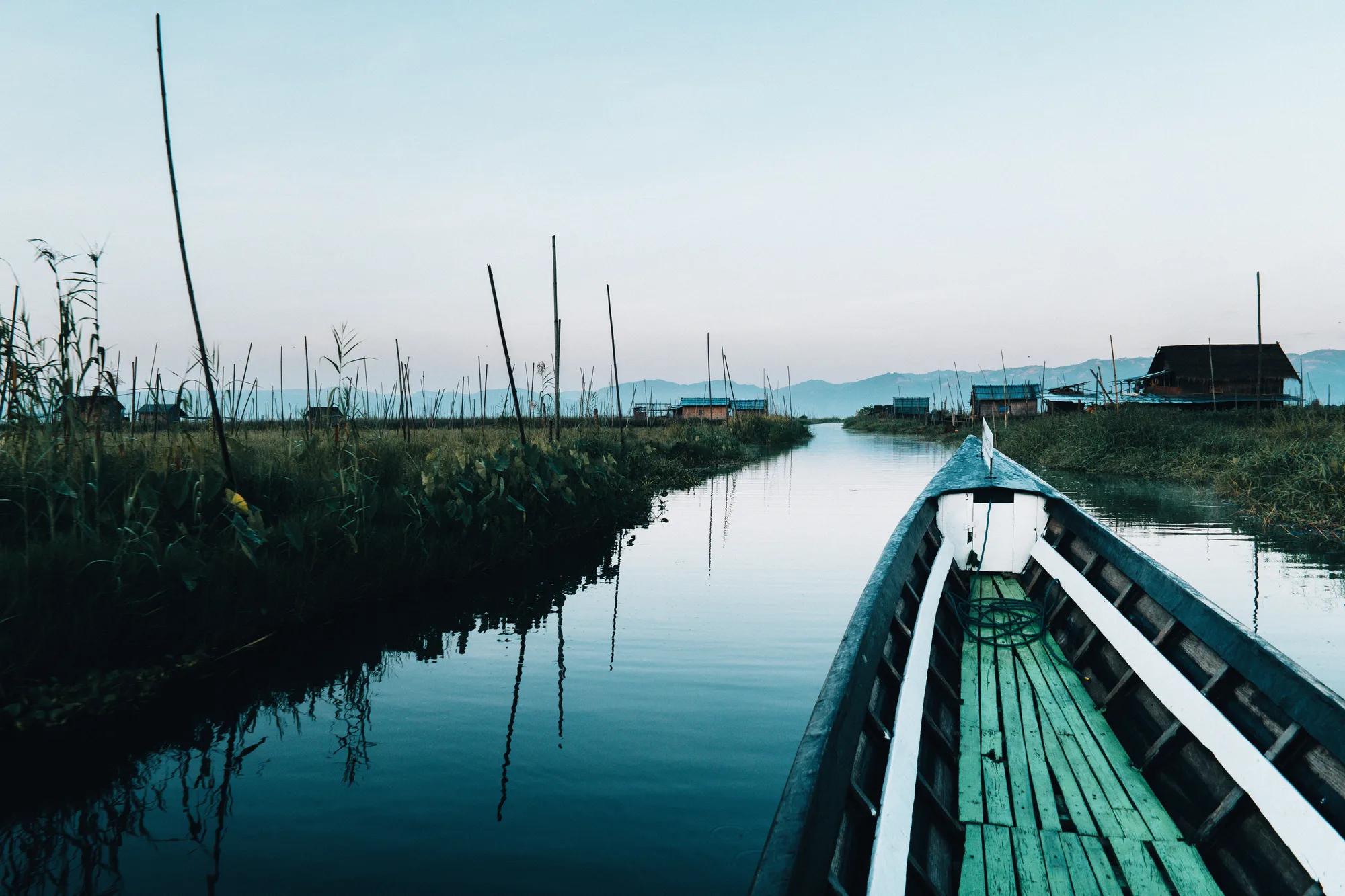 A long boat moving down a still canal past stilt houses in the blue light before sunrise on Inle Lake