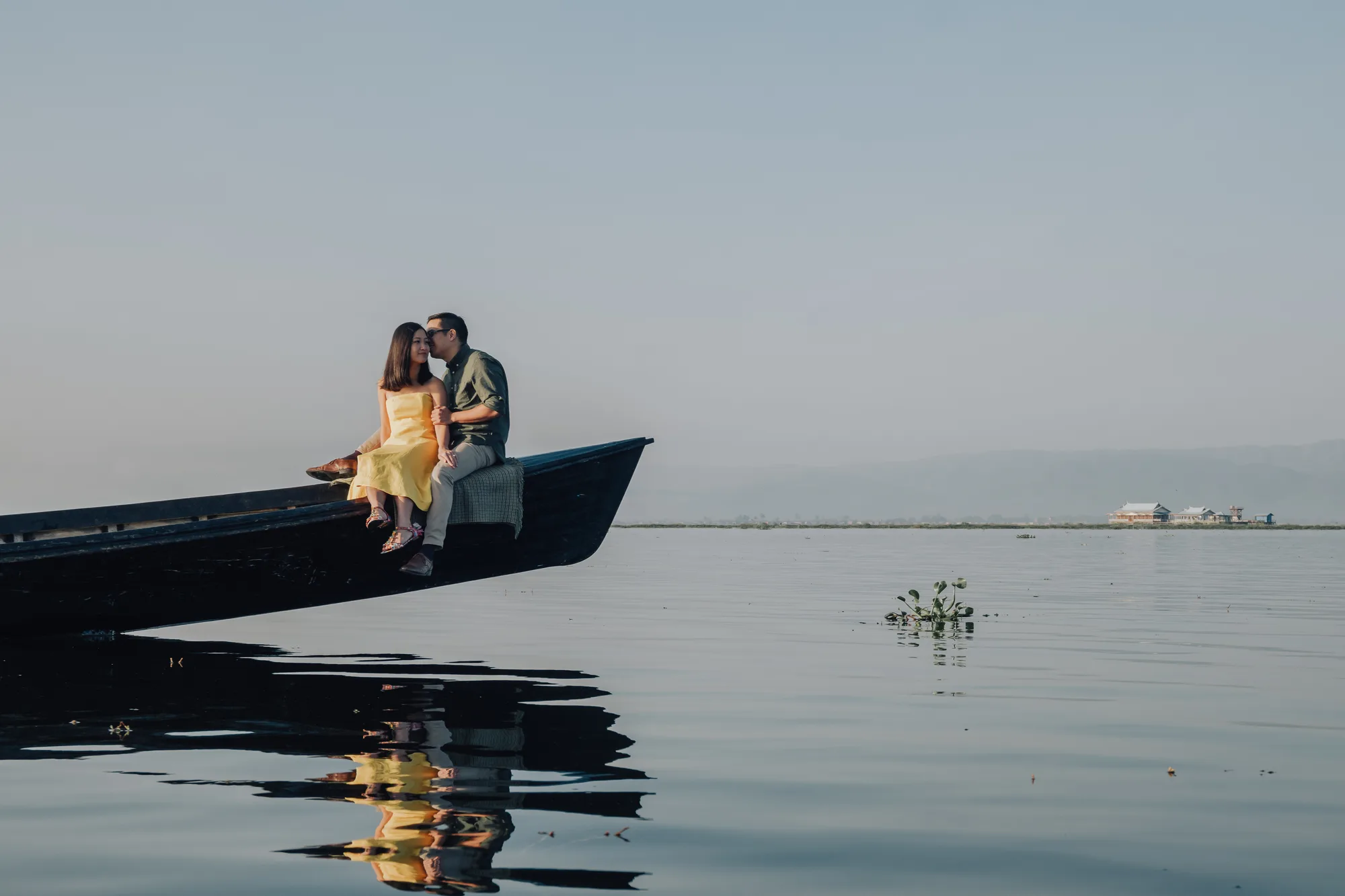 A couple shares a tender moment on the edge of a boat in a serene lake setting under soft evening light.