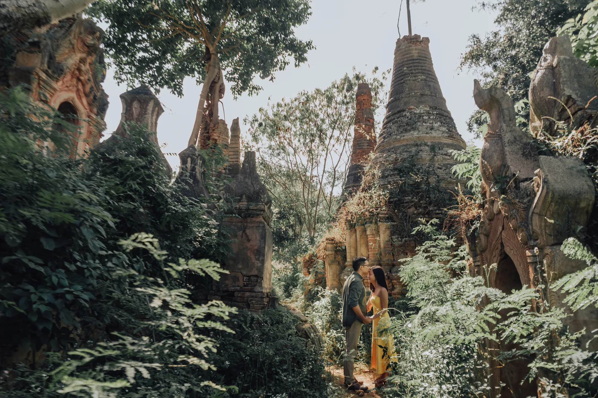 A couple stands amidst ancient temple ruins in a lush forest setting under soft natural light.