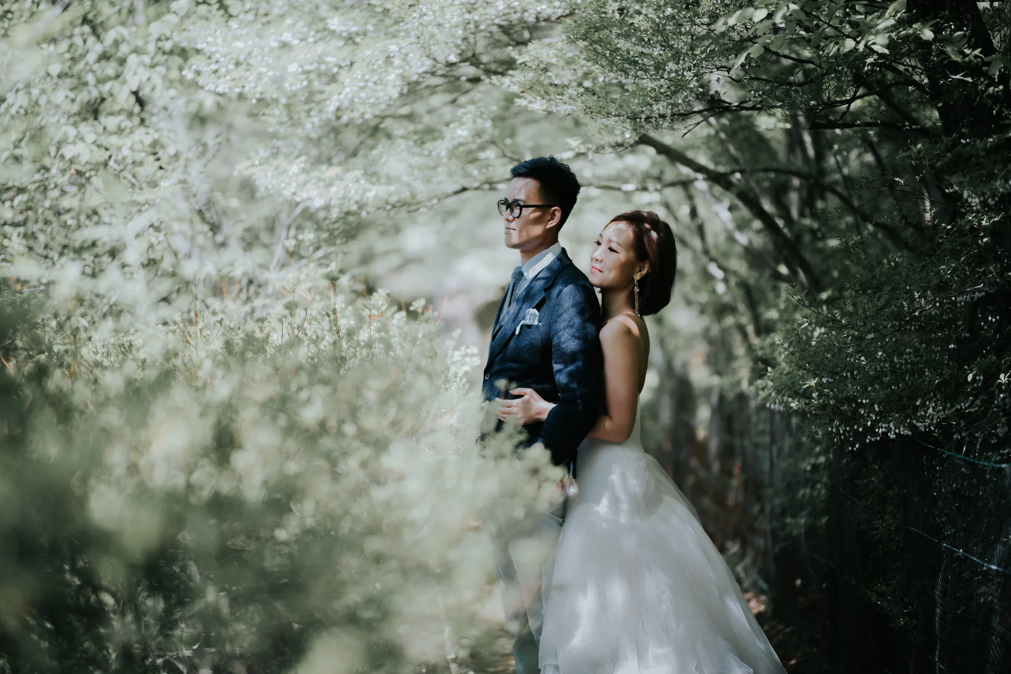 A couple stands close together amidst blooming white flowers in a serene forest setting