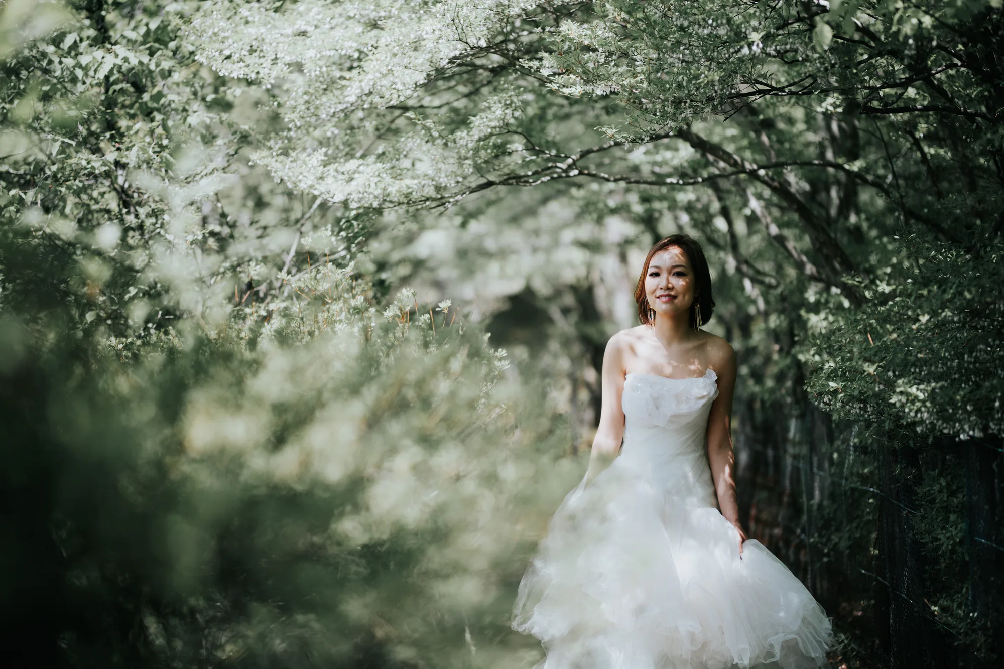 A bride in a white gown stands amidst blooming trees, bathed in soft, natural light filtering through the foliage.