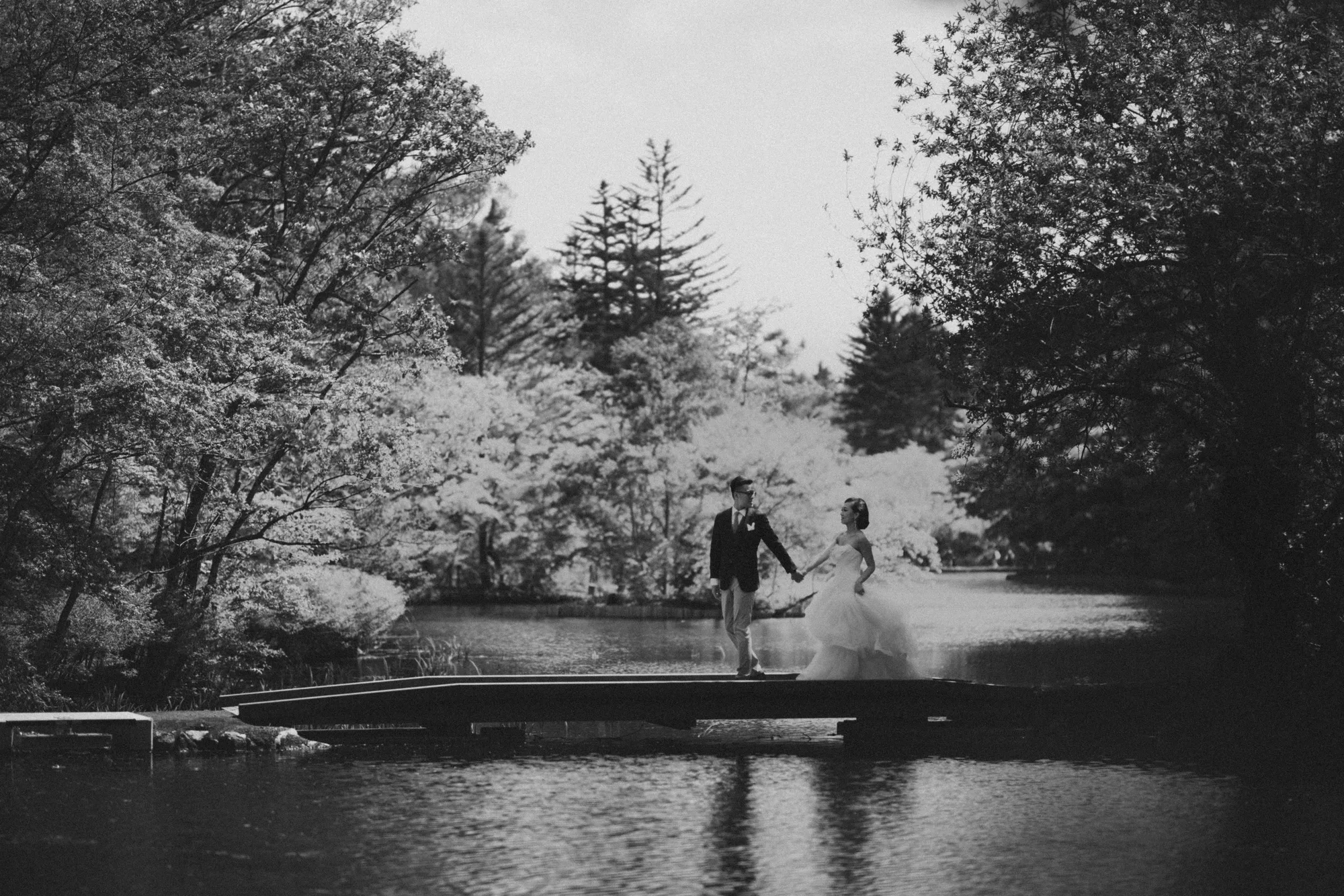 A bride and groom walk hand in hand across a bridge over a serene pond surrounded by lush greenery under soft natural light.