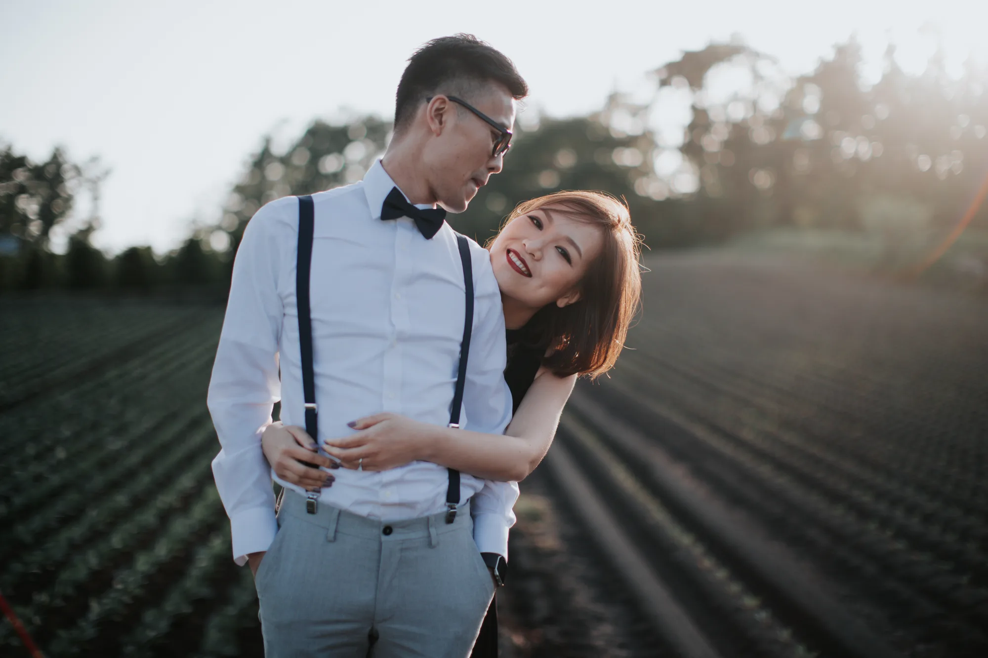 A couple stands close in a serene field at sunset, bathed in warm light