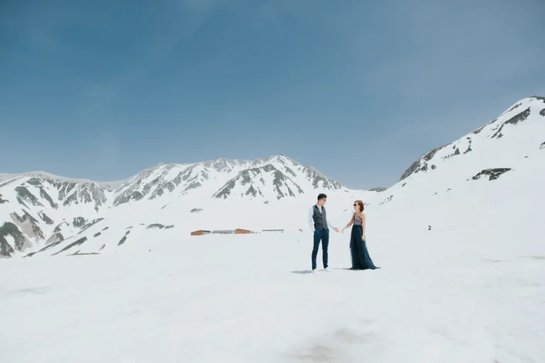 A couple stands hand-in-hand in a snowy mountain landscape under clear blue skies