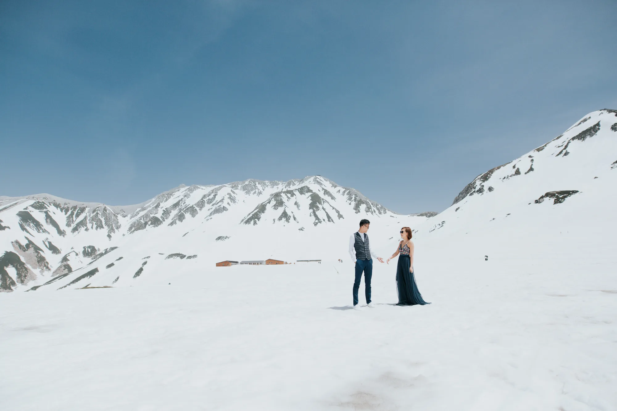 A couple stands hand-in-hand in a snowy mountain landscape under clear blue skies
