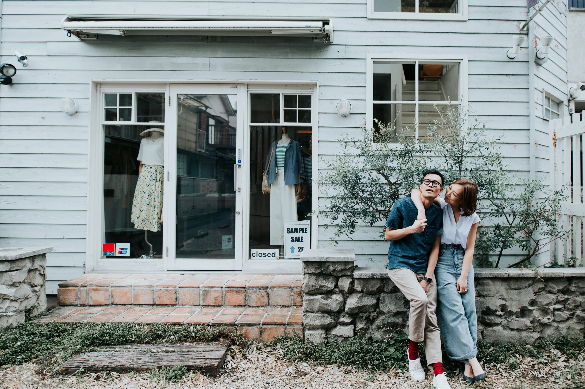 A couple leans affectionately against a stone wall in front of a quaint shop window during the day