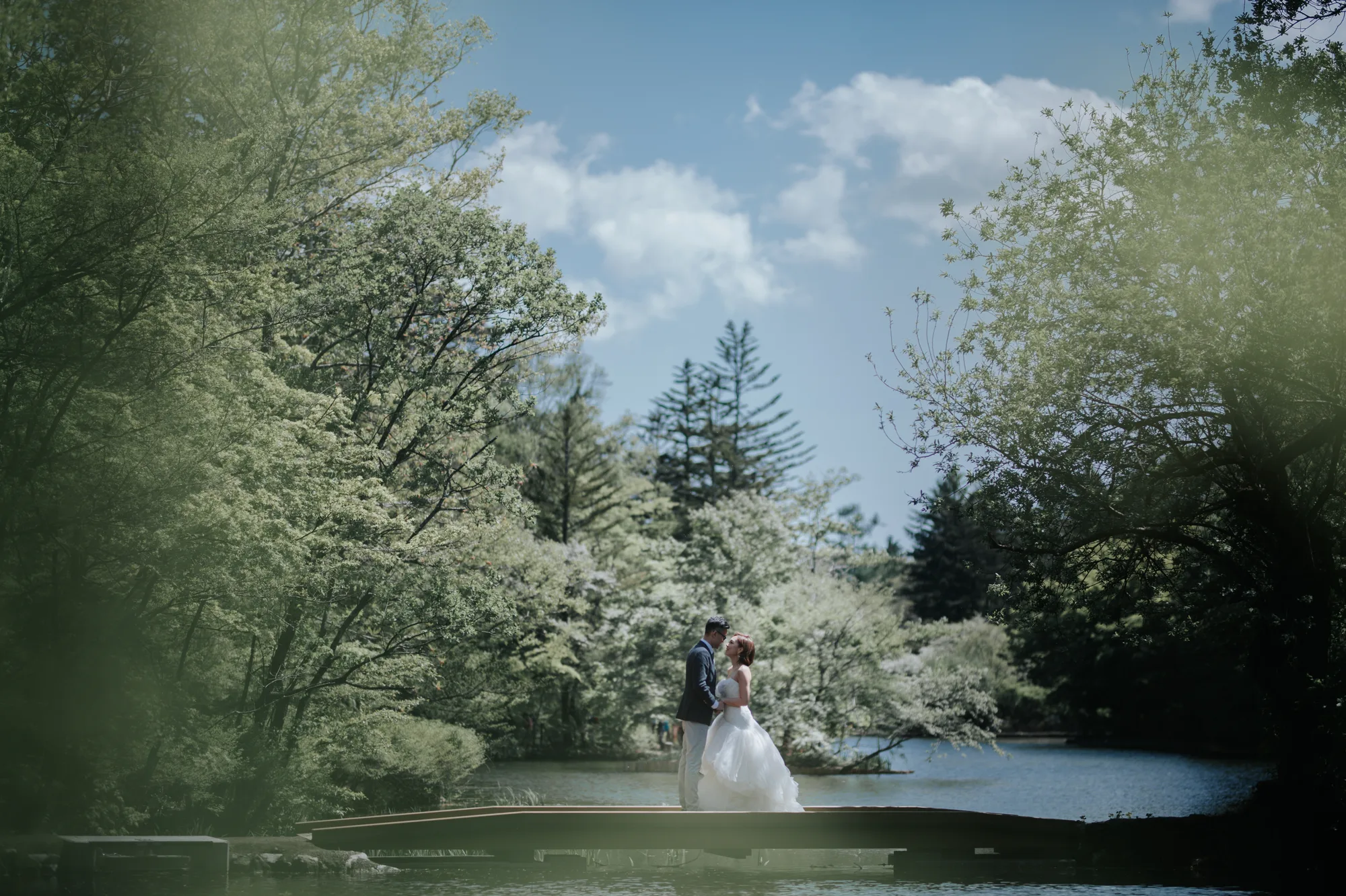 A bride and groom stand on a bridge in a serene park setting under bright sunlight