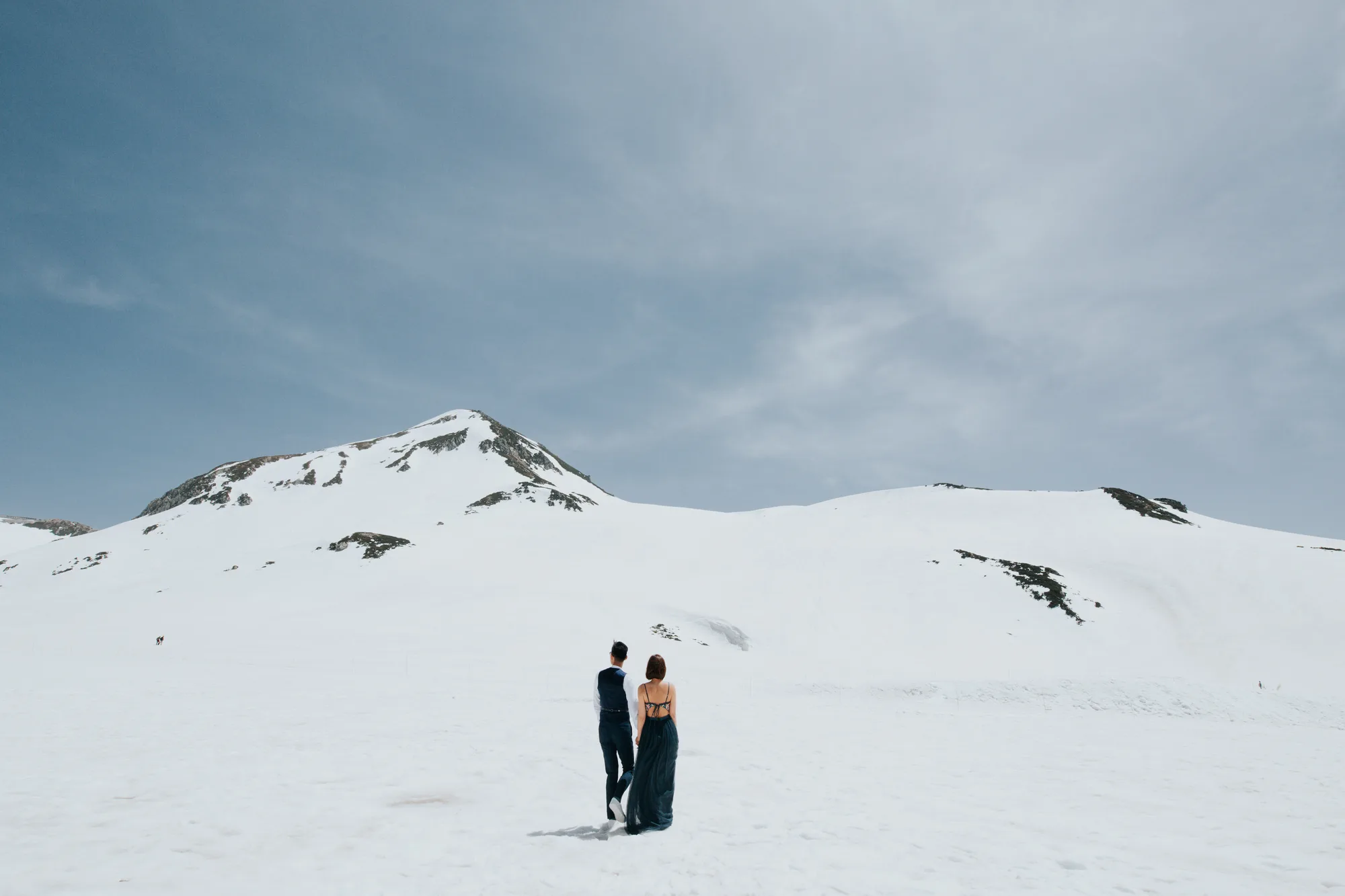 A couple walks hand in hand across a snowy mountain landscape under a bright blue sky.