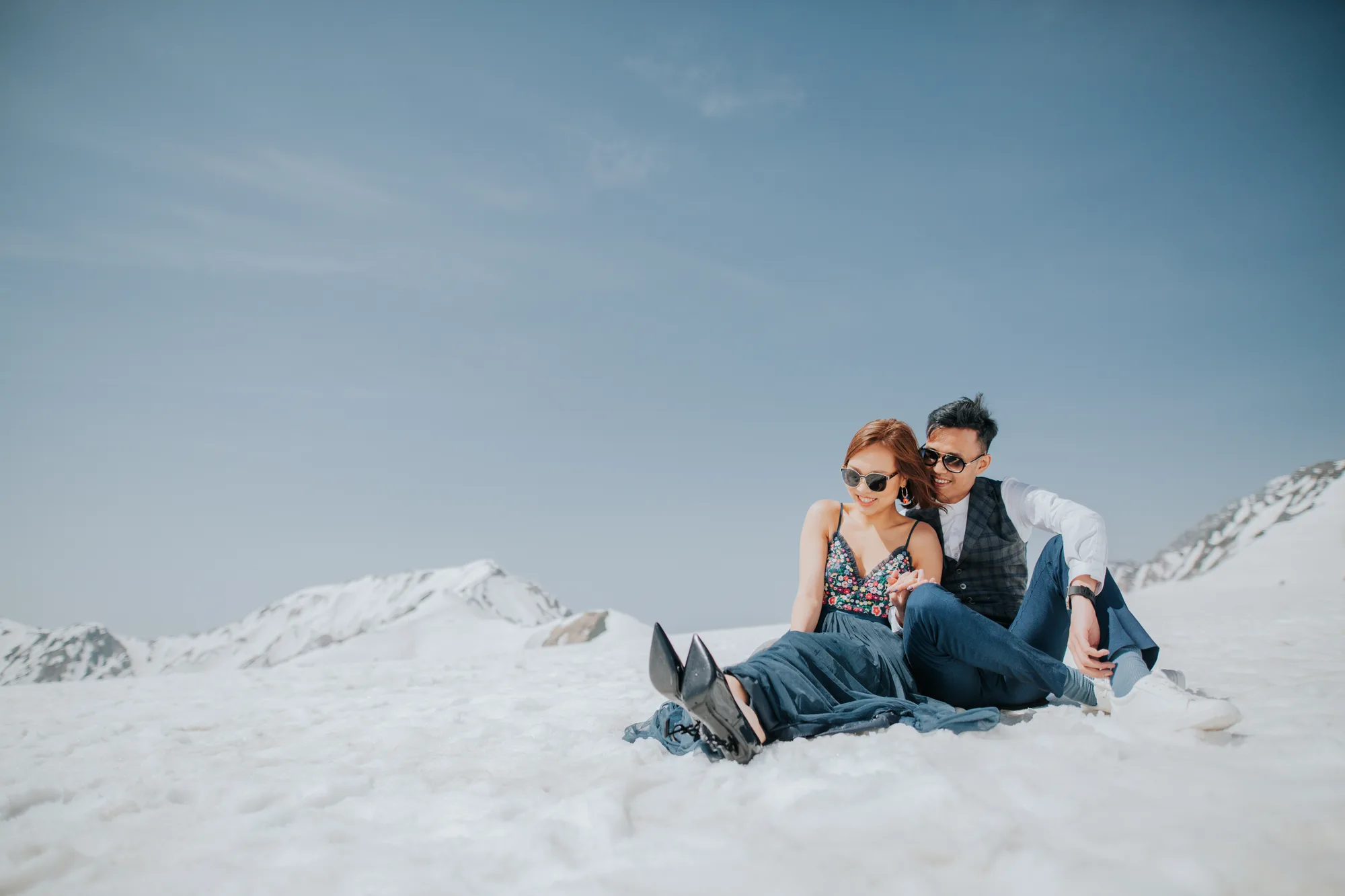 A couple sits on a snowy mountain peak under clear blue skies