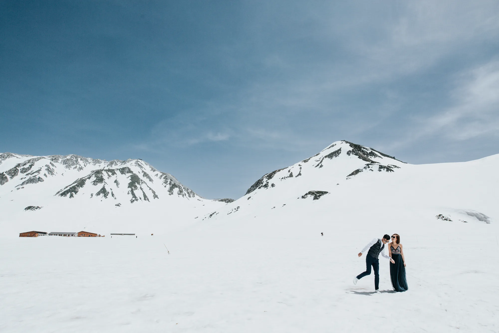 A couple in formal attire enjoys a playful moment on a snowy mountain landscape under bright sunlight.