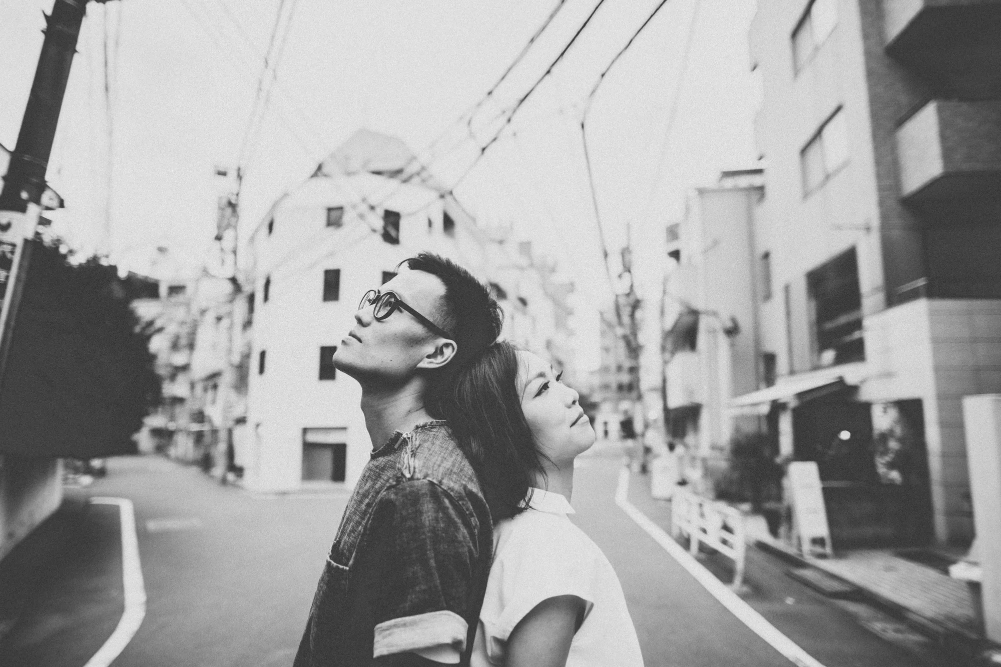 A couple stands back-to-back on a quiet urban street under overcast skies
